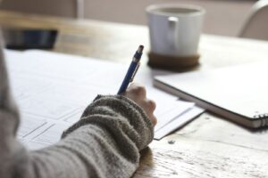 A hand writing in a notebook on a wooden desk with a coffee cup — turning a private AI journey into a public playbook, blog, and book.