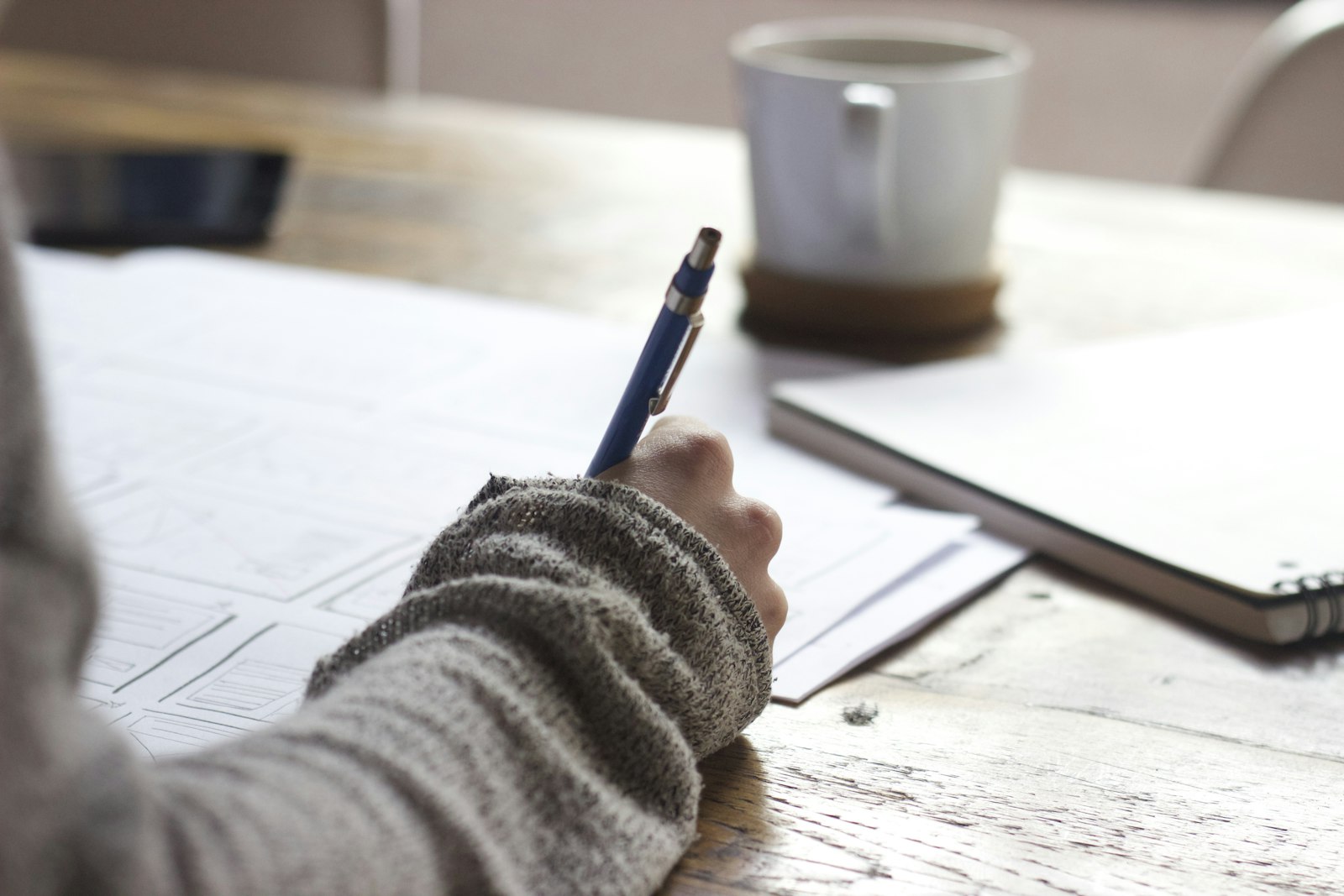 A hand writing in a notebook on a wooden desk.